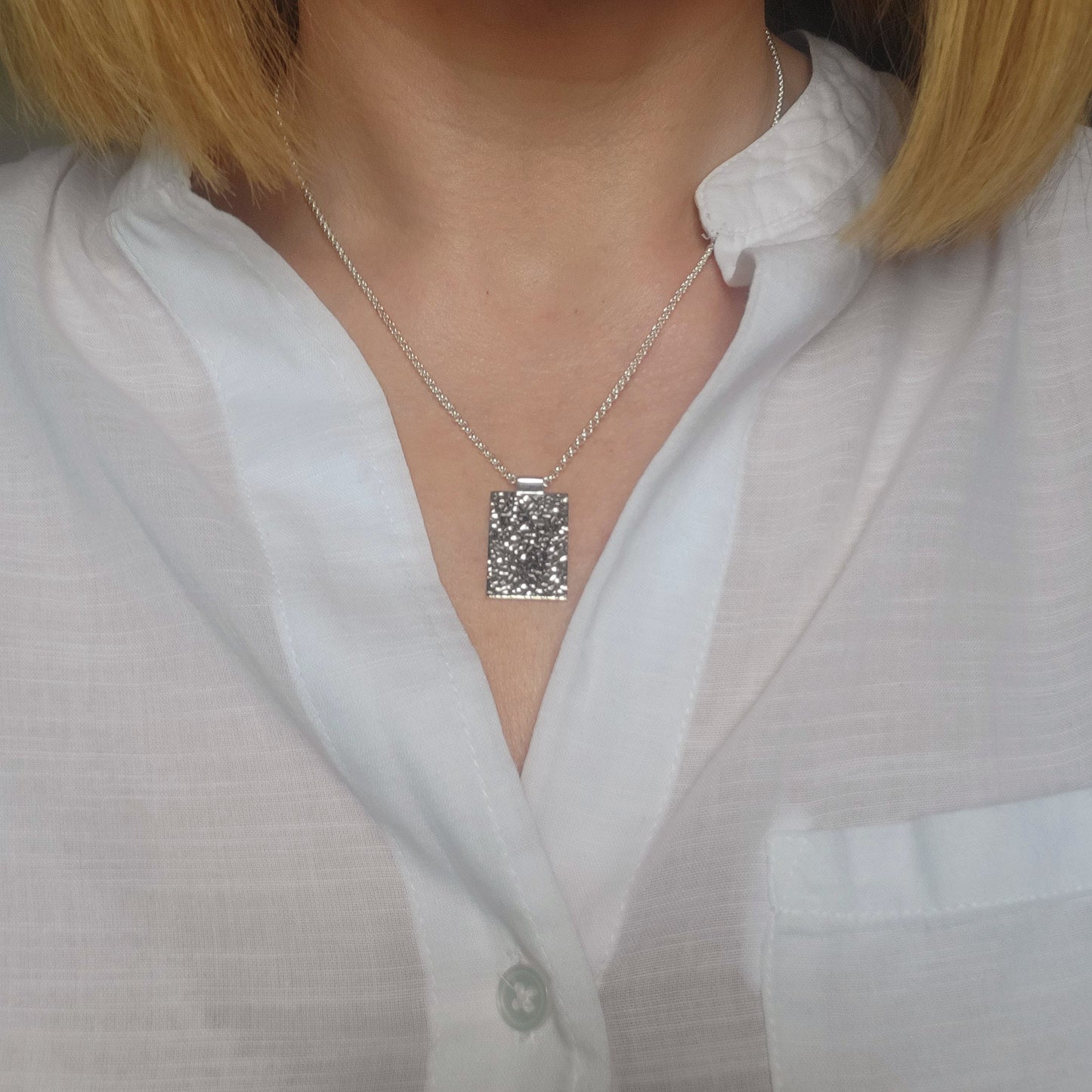 Silver necklace with a rectangular hammered pendant worn by a person in a white shirt.